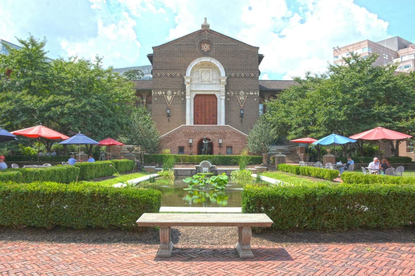Penn Museum A courtyard with a central pond and lily pads, surrounded by hedges, outdoor seating with colorful umbrellas, and a brick building in the background.