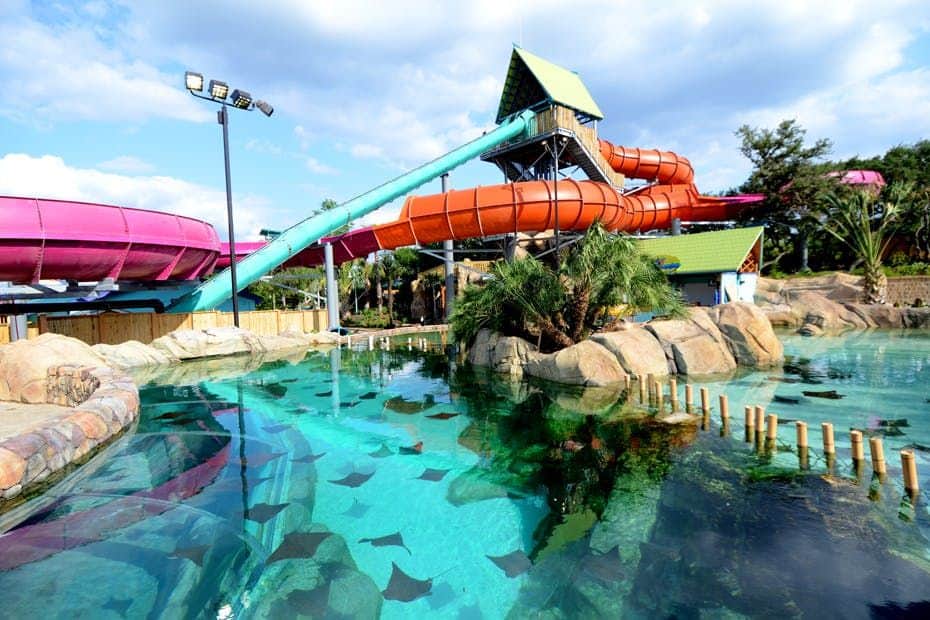 Aquatica San Antonio Water park with colorful slides, clear pools with stingrays, rocky landscaping, and palm trees under a partly cloudy sky.