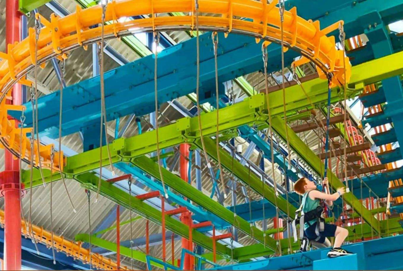 LiggettVille Adventure Center A young boy climbs a colorful indoor ropes course with blue, green, orange, and red structures and safety harnesses.