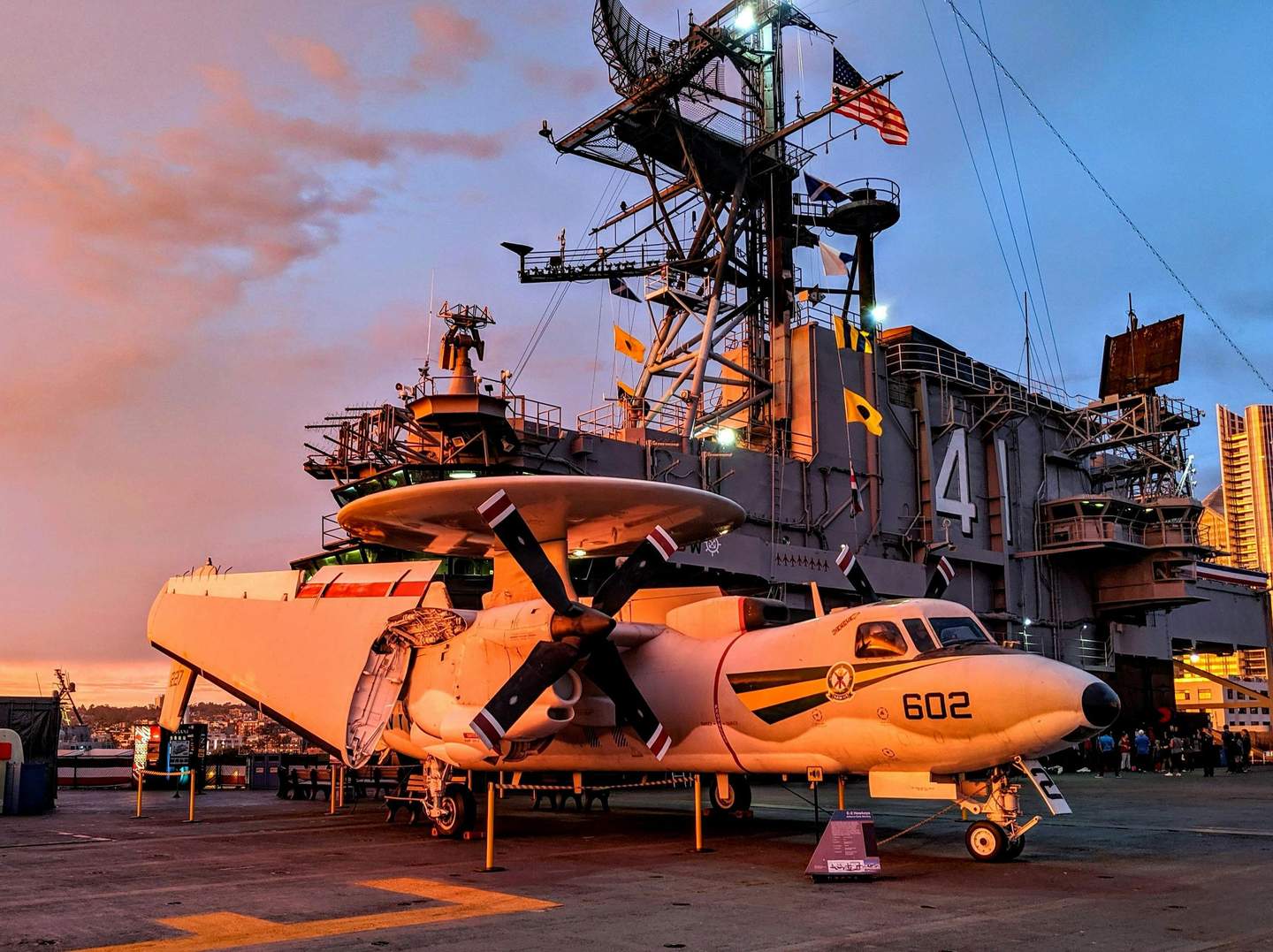 USS Midway Museum A white propeller aircraft marked "602" positioned on the deck of a naval ship at sunset, with an American flag flying.