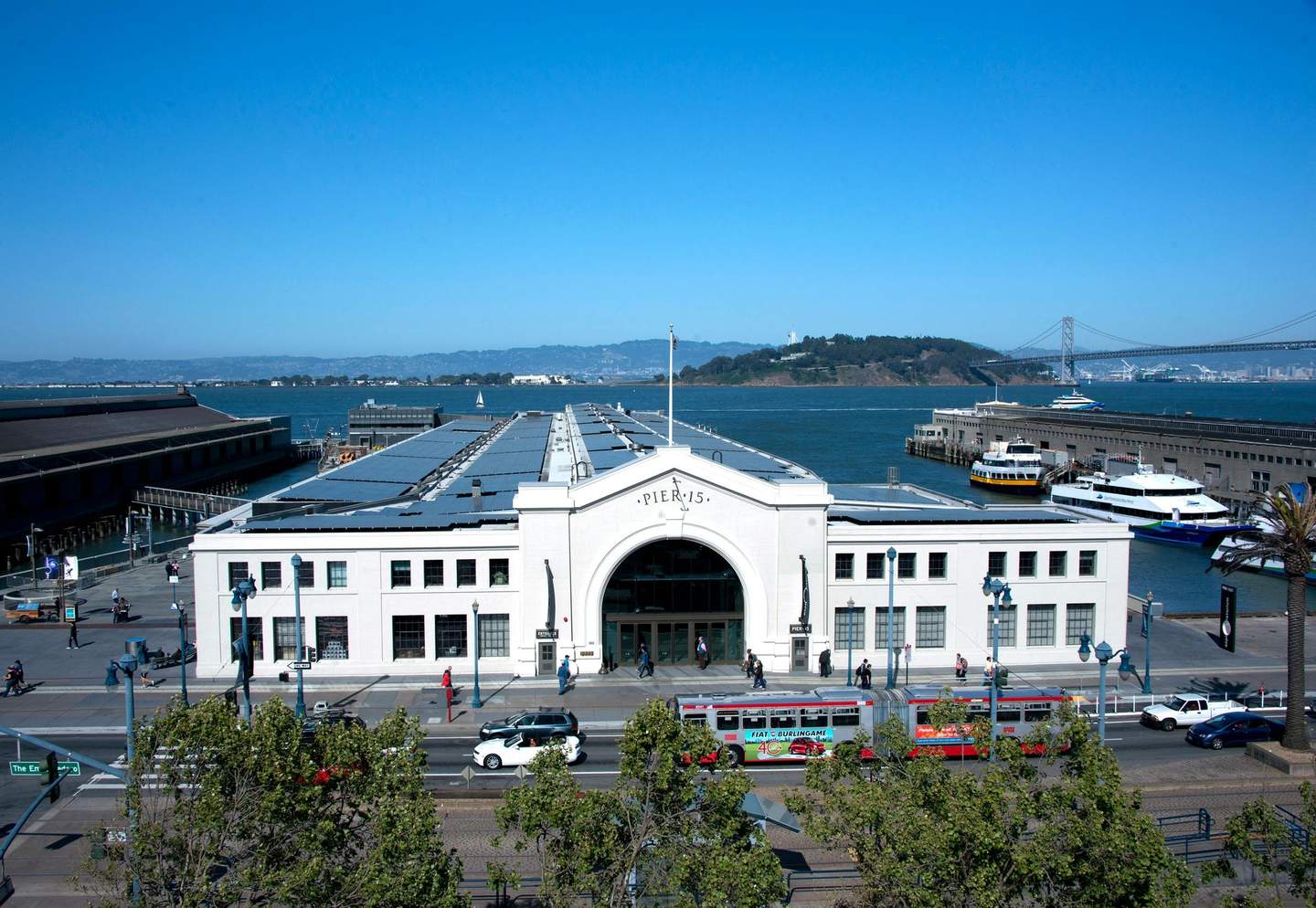 Exploratorium The image shows Pier 15 in San Francisco, featuring a large white building, a bay, boats, people walking, and the Bay Bridge in the background.