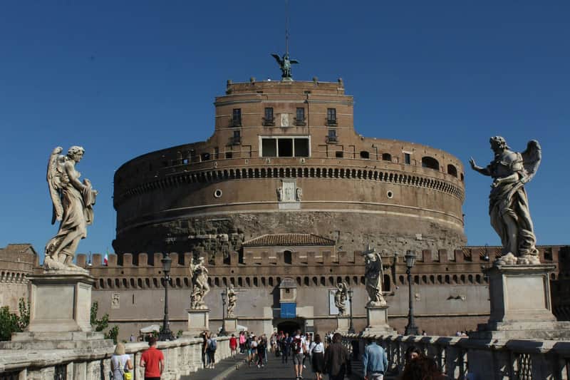 Castel Sant'Angelo: Skip The Line Ticket + Guided Tour Large, circular brick fortress with a statue on top, viewed from a crowded pedestrian bridge adorned with statues under a clear blue sky.