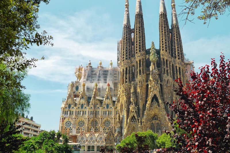 Sagrada Familia: Skip the line-ticket + Rondleiding met gids Een grote, ingewikkelde kathedraal in gotische stijl met meerdere torenspitsen onder een blauwe hemel, omringd door groen en bomen.