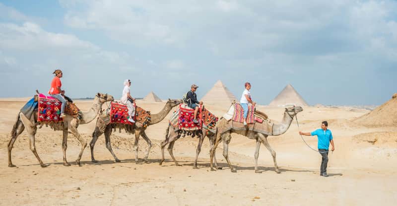Quatre personnes montent à dos de chameau sous la conduite d'un guide dans un désert avec des pyramides en arrière-plan, sous un ciel partiellement nuageux.