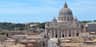 View of St. Peter's Basilica with its large dome, surrounded by rooftops and trees under a clear blue sky.