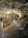 Une grotte avec de nombreuses stalactites suspendues au plafond et des stalagmites s'élevant du sol, avec une passerelle en arrière-plan.