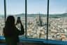 A person photographing the cityscape of Barcelona through a large window, with the Sagrada Família visible in the distance.