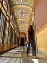 Une femme marche dans un couloir très éclairé, doté de grandes fenêtres et d'un plafond richement décoré et orné.