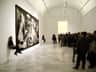 Visitors observe Picasso's "Guernica" painting in a museum. Guides sit on chairs near the artwork.