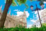 Gothic cathedral with intricate spires, a tall bell tower, nearby street lamp, lush greenery, and a bright blue sky with clouds.