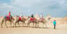 Planalto de Gizé Quatro pessoas montam camelos conduzidos por um guia num deserto com pirâmides ao fundo, sob um céu parcialmente nublado.
